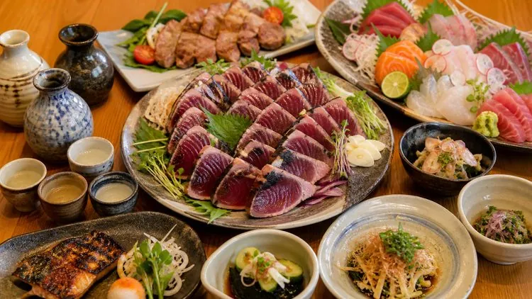 夕食会のイメージ——カツオのたたきや刺身、日本酒が並ぶ宴のテーブル（イメージ画像）
